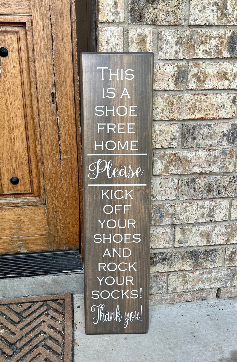 May include: A wooden sign with the text "This is a shoe free home. Please kick off your shoes and rock your socks! Thank you!" in white lettering.