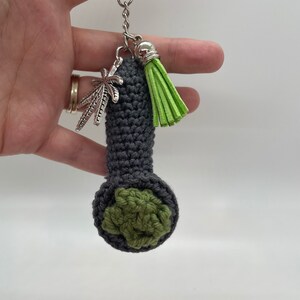 May include: A gray crocheted pipe keychain with a green crocheted bowl and a silver marijuana leaf charm. The keychain also has a green tassel.