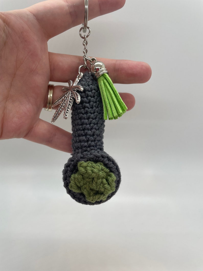May include: A gray crocheted pipe keychain with a green crocheted bowl and a silver marijuana leaf charm. The keychain also has a green tassel.