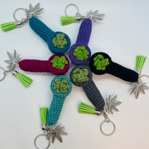 May include: Five crocheted pipe keychains with green marijuana leaf charms. Each keychain is a different color: teal, purple, pink, gray, and black.
