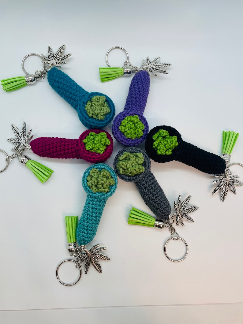 May include: Five crocheted pipe keychains with green marijuana leaf charms. Each keychain is a different color: teal, purple, pink, gray, and black.