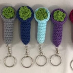 May include: Six crochet keychains in a row, each featuring a different color: red, gray, navy blue, light blue, purple, and magenta. Each keychain has a bowl shape filled with green crochet, and a silver keyring.