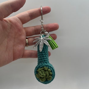 May include: A teal crocheted pipe keychain with a green crocheted bowl and a silver marijuana leaf charm. The keychain also has a green tassel.