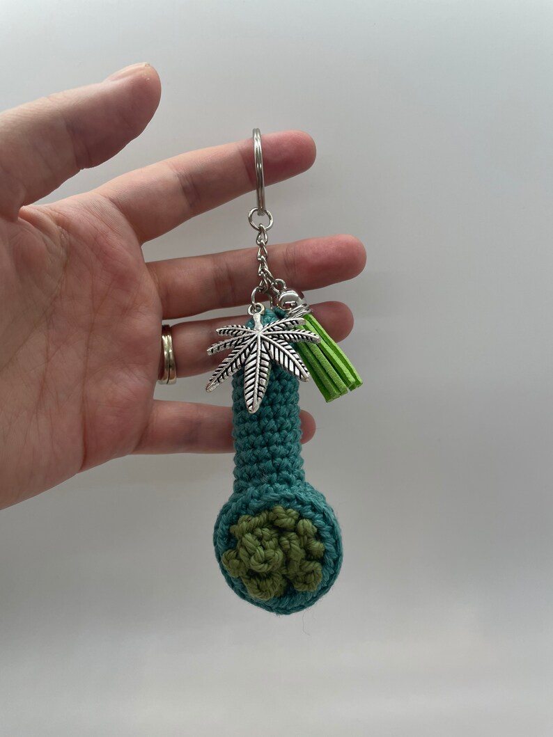 May include: A teal crocheted pipe keychain with a green crocheted bowl and a silver marijuana leaf charm. The keychain also has a green tassel.