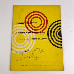 May include: A yellow programme booklet for the musical "Annie Get Your Gun" starring Mary Martin and John Raitt. The cover features a stylised illustration of a woman riding a horse, surrounded by concentric circles in red, white and black.
