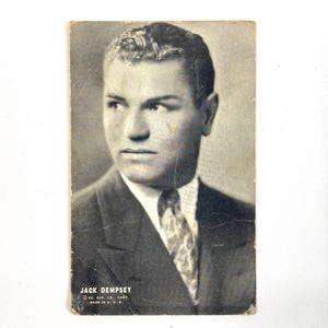 May include: A vintage black and white photograph of Jack Dempsey. He is wearing a suit and tie. The photograph has the text "JACK DEMPSEY" and "EX. SUP. CO., CHGO. MADE IN U. S. A." at the bottom.
