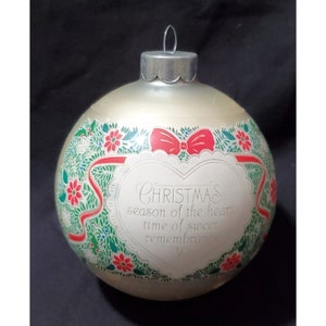 May include: A white glass Christmas ornament with a red bow and green holly leaves. The ornament has a heart-shaped label with the text "Christmas season of the heart time of sweet remembrance".
