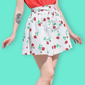 Puede incluir: Una falda blanca con rosas rojas y lunares blancos. La falda tiene una cintura alta y está ajustada a las caderas. La falda la lleva una persona con un tatuaje en el brazo.