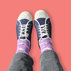 May include: A pair of blue canvas trainers with white laces and a white toe cap. The trainers are worn with purple socks featuring a white dolphin pattern.