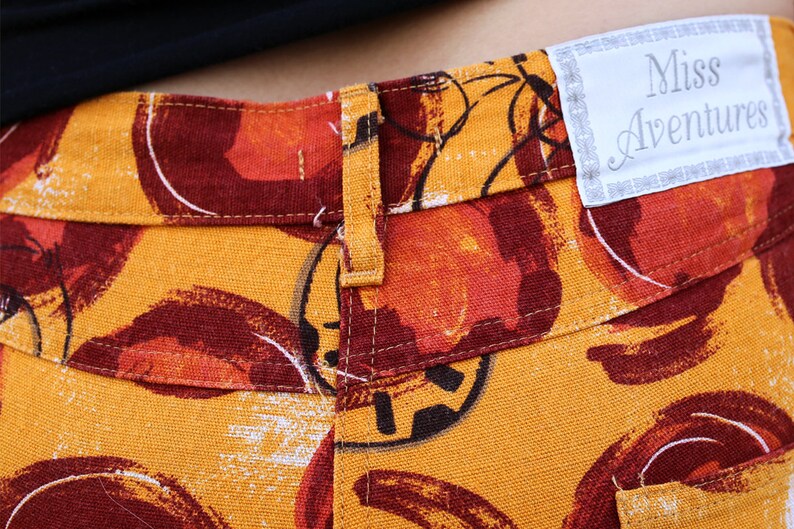 Puede incluir: Pantal&oacute;n amarillo con estampado abstracto rojo y naranja. El pantal&oacute;n tiene cierre de bot&oacute;n y una etiqueta que dice "Miss Adventures".