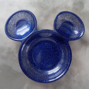 May include: A blue glitter resin dish shaped like a mouse head, featuring two circular ears. The dish has a glossy finish with silver glitter. The ears are a slightly lighter shade of blue than the main body of the dish.