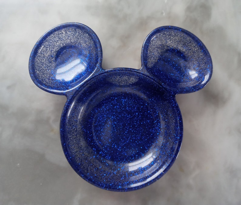 May include: A blue glitter resin dish shaped like a mouse head, featuring two circular ears. The dish has a glossy finish with silver glitter. The ears are a slightly lighter shade of blue than the main body of the dish.