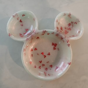 May include: A white, Mickey Mouse-shaped trinket dish with two small ear-shaped dishes and a larger circular dish. The dish is speckled with red Mickey Mouse-shaped confetti and has a light green hue.