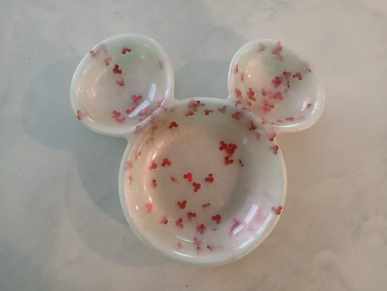 May include: A white, Mickey Mouse-shaped trinket dish with two small ear-shaped dishes and a larger circular dish. The dish is speckled with red Mickey Mouse-shaped confetti and has a light green hue.
