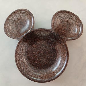 May include: A brown, glitter-covered dish shaped like Mickey Mouse's head. It features a large circular bowl and two smaller, rounded ear-shaped sections. The surface has a glossy finish with flecks of glitter.