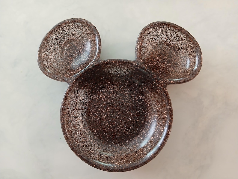 May include: A brown, glitter-covered dish shaped like Mickey Mouse's head. It features a large circular bowl and two smaller, rounded ear-shaped sections. The surface has a glossy finish with flecks of glitter.
