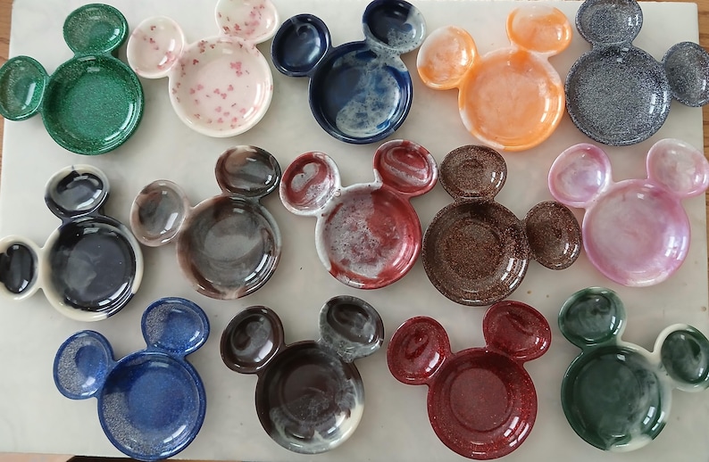 May include: A selection of ceramic dishes, each shaped like Mickey Mouse's head. The dishes are in a variety of colours, such as green, pink, blue, orange, and brown. Each has a glossy finish and features a small ear-shaped dish on either side of the main bowl.