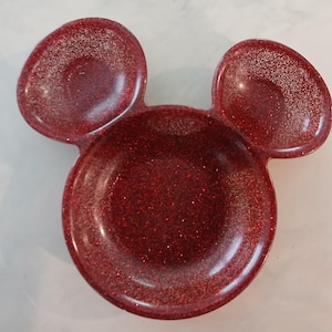 May include: A red glitter trinket dish shaped like Mickey Mouse's head. The dish features a circular base with two rounded ear-shaped sections. The shiny dish is made of a reflective material.