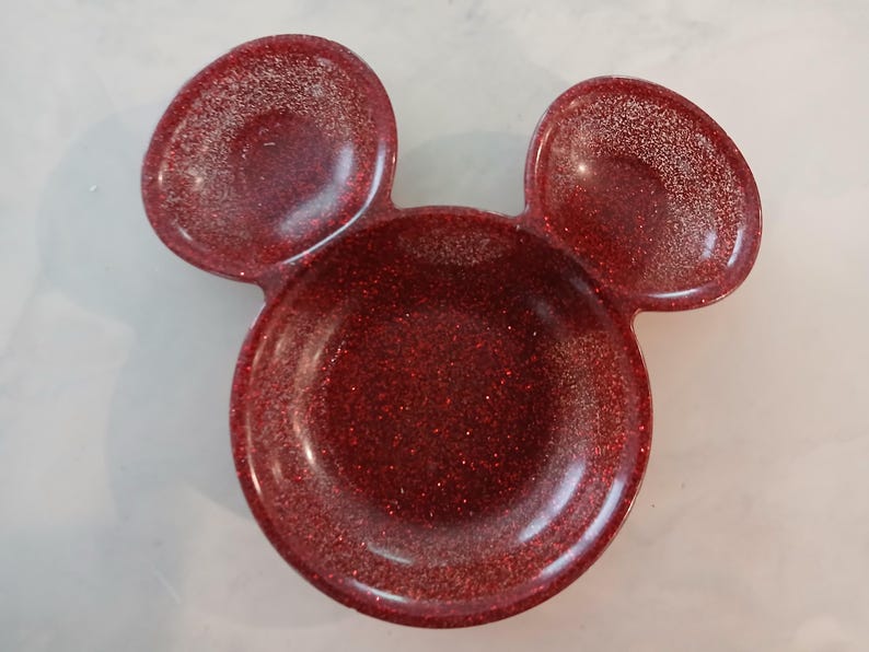 May include: A red glitter trinket dish shaped like Mickey Mouse's head. The dish features a circular base with two rounded ear-shaped sections. The shiny dish is made of a reflective material.