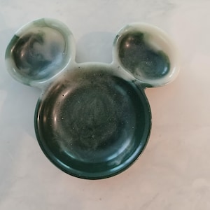 May include: A resin trinket dish shaped like Mickey Mouse's head, with a dark green and white marbled pattern. The dish has a circular base and two ear-shaped sections. The measurements are approximately 15 cm wide and 10 cm high.