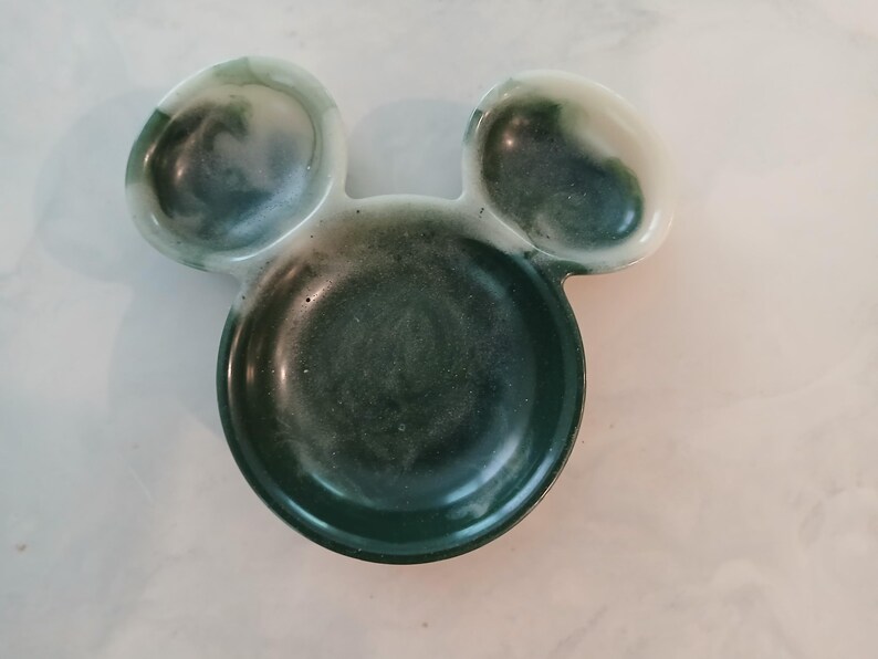 May include: A resin trinket dish shaped like Mickey Mouse's head, with a dark green and white marbled pattern. The dish has a circular base and two ear-shaped sections. The measurements are approximately 15 cm wide and 10 cm high.