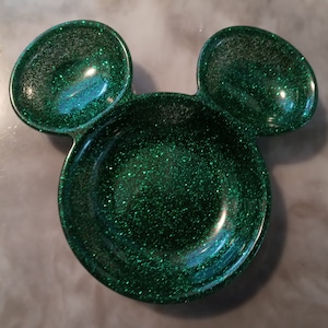 May include: A green glitter resin dish shaped like Mickey Mouse's head. It features a large circular bowl and two smaller ear-shaped bowls. The glitter catches the light, giving it a sparkly appearance.