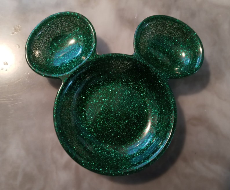 May include: A green glitter resin dish shaped like Mickey Mouse's head. It features a large circular bowl and two smaller ear-shaped bowls. The glitter catches the light, giving it a sparkly appearance.