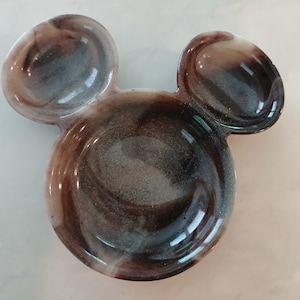 May include: A decorative dish shaped like Mickey Mouse's head. It features a marbled design in shades of brown, grey, and white. The dish has two rounded ears and a circular base, with a glossy finish.