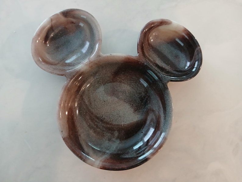 May include: A decorative dish shaped like Mickey Mouse's head. It features a marbled design in shades of brown, grey, and white. The dish has two rounded ears and a circular base, with a glossy finish.
