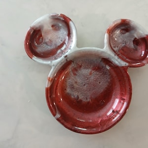 May include: A red, white, and clear resin dish shaped like Mickey Mouse's head. The dish has a glossy finish with a circular base and two smaller circular ears. The red colour has a marbled effect.