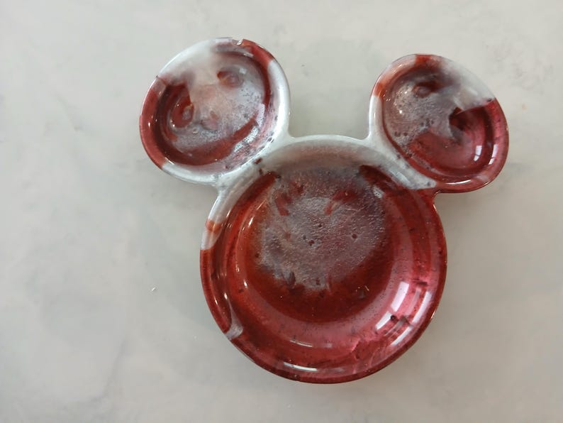 May include: A red, white, and clear resin dish shaped like Mickey Mouse's head. The dish has a glossy finish with a circular base and two smaller circular ears. The red colour has a marbled effect.