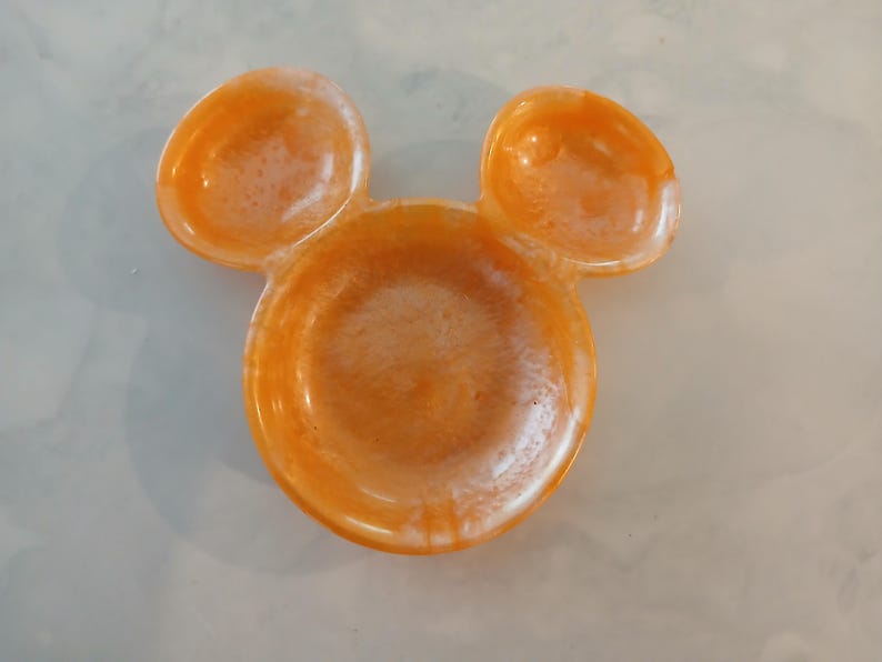 May include: Orange resin dish shaped like a mouse head, with two ear-shaped dishes above the main bowl. The dish has a glossy finish and a marbled effect.