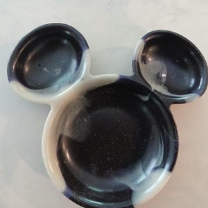May include: A decorative dish in the shape of Mickey Mouse's head. The dish is a glossy resin with a dark blue and white swirl pattern. The dish has two smaller ear-shaped sections and a larger circular section.