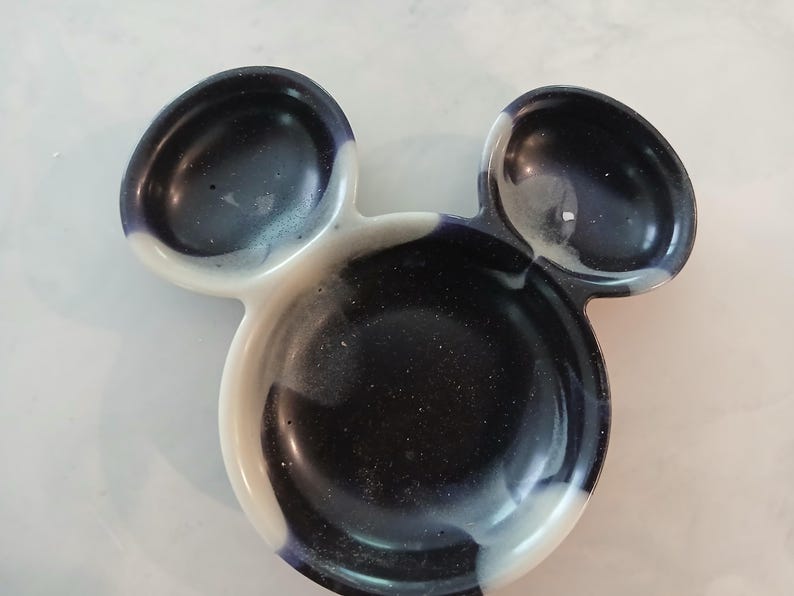 May include: A decorative dish in the shape of Mickey Mouse's head. The dish is a glossy resin with a dark blue and white swirl pattern. The dish has two smaller ear-shaped sections and a larger circular section.