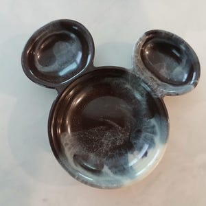 May include: A decorative tray in the shape of Mickey Mouse's head. The tray is black with white and grey swirls, and features two small ear-shaped compartments and a larger circular compartment for holding items.