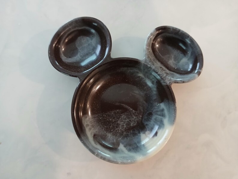 May include: A decorative tray in the shape of Mickey Mouse's head. The tray is black with white and grey swirls, and features two small ear-shaped compartments and a larger circular compartment for holding items.