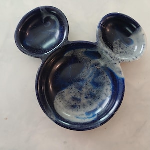 May include: A ceramic dish shaped like Mickey Mouse's head. The dish is a deep blue with white and light blue accents. It has three sections: two small ear-shaped compartments and a larger central section.