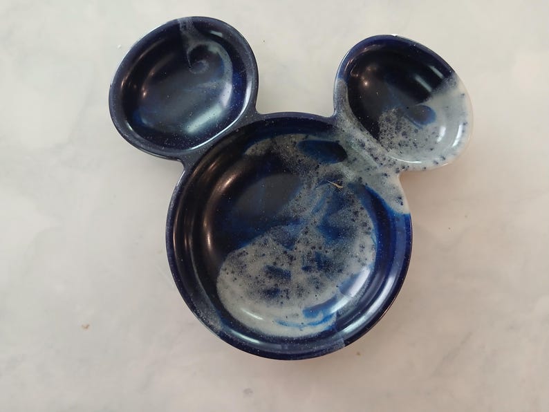 May include: A ceramic dish shaped like Mickey Mouse's head. The dish is a deep blue with white and light blue accents. It has three sections: two small ear-shaped compartments and a larger central section.