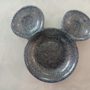 May include: A dark grey, glitter-covered dish in the shape of Mickey Mouse's head. It features a large circular base with two smaller, rounded sections above, mimicking ears. The surface is covered in sparkling glitter.