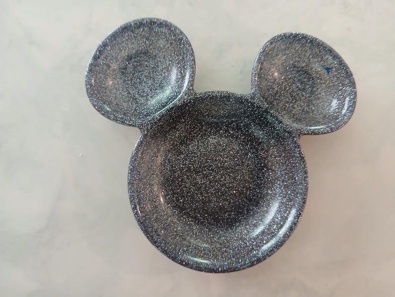 May include: A dark grey, glitter-covered dish in the shape of Mickey Mouse's head. It features a large circular base with two smaller, rounded sections above, mimicking ears. The surface is covered in sparkling glitter.