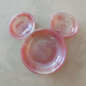 May include: A pink and white resin trinket dish shaped like a mouse head. The dish features a circular base with two smaller, rounded ear-shaped dishes above. The resin has a glossy finish.