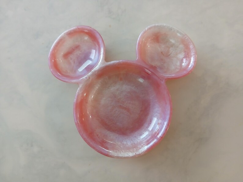 May include: A pink and white resin trinket dish shaped like a mouse head. The dish features a circular base with two smaller, rounded ear-shaped dishes above. The resin has a glossy finish.