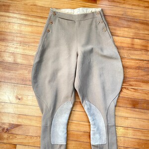 May include: Tan riding breeches with suede knee patches and button details. The breeches have a high waist and flared thighs, with a tailored fit. The legs are straight with a small slit at the ankle.