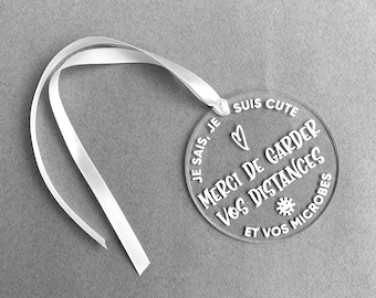 May include: A white acrylic circle with a white ribbon attached. The circle has the text "Je sais, je suis cute" and "Merci de garder vos distances et vos microbes" in French.