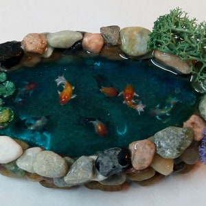 May include: A miniature pond with a blue resin water surface, surrounded by small stones and featuring several orange and white goldfish. The pond is decorated with green and pink lily pads and two small green and purple plants.