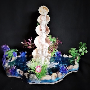 May include: A tabletop fountain with a cascading water feature. The fountain is made of clear plastic and has a layered design. The base of the fountain is surrounded by artificial plants and rocks.