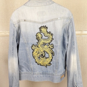 Puede incluir: Chaqueta vaquera azul claro con un diseño de dragón dorado bordado en la espalda. La chaqueta tiene cuello clásico, puños abotonados y un lavado descolorido. Una prenda de vestir exterior elegante.