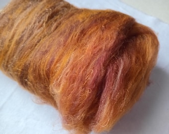 May include: A bundle of unspun fiber roving featuring a variegated color palette of warm orange, deep rust, and soft golden yellow tones. The texture is fine, airy, and slightly fuzzy, with long, flowing strands arranged in a dense, cylindrical shape.