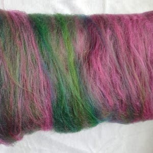 May include: A rectangular batt of unspun fiber featuring a variegated blend of vibrant magenta, deep forest green, and dark charcoal gray tones. The soft, airy material displays a long, fibrous texture with overlapping color gradients throughout.
