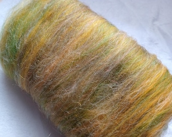 May include: A cylindrical roll of unspun wool roving featuring a variegated blend of fibers in shades of golden yellow, soft orange, and muted olive green. The texture is fine, airy, and slightly fuzzy, with individual strands visible throughout the roll.